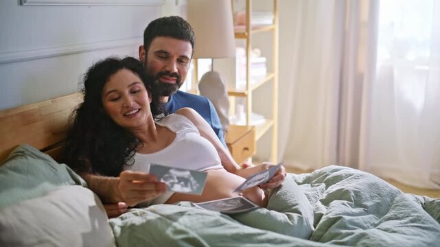 Happy couple looking ultrasound photo bed. Pregnant wife husband bonding home.