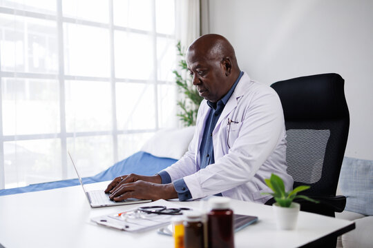 Senior african american doctor wearing a lab coat and using a laptop computer at his desk in a clinic, managing patient records and modern healthcare administration