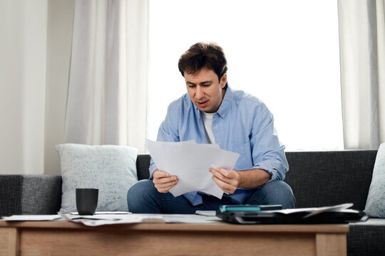 Stressed man reading overdue utility bills at home. He is worried about rising household expenses, inflation, and financial debt during an economic crisis.