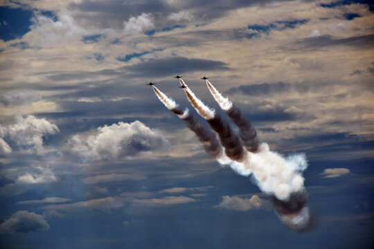 Fighter jets perform a precision formation flight leaving dramatic smoke trails across a cloudy sky.