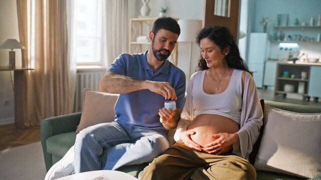 Man applying moisturizer cream pregnant belly. Loving couple bonding home sofa.
