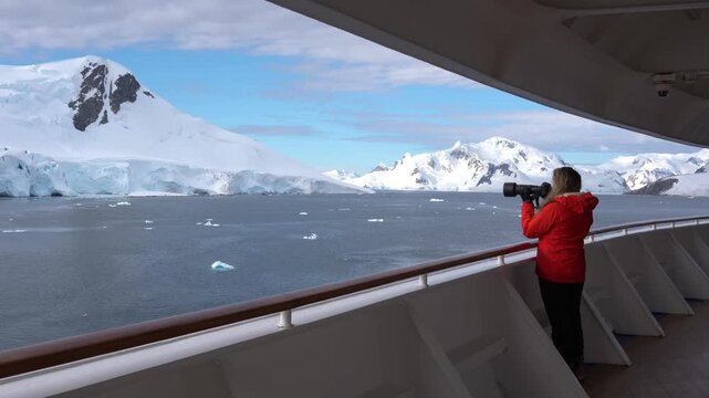 Female Photographer Antarctica Cruise Ship Passenger in Orange Coat on Deck. Epic Adventure Travel Snow Covered Mountains Telephoto Lens Professional
