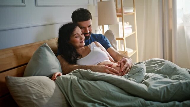 Couple relaxing bed cozy morning. Man cradling pregnant wife belly bedroom.
