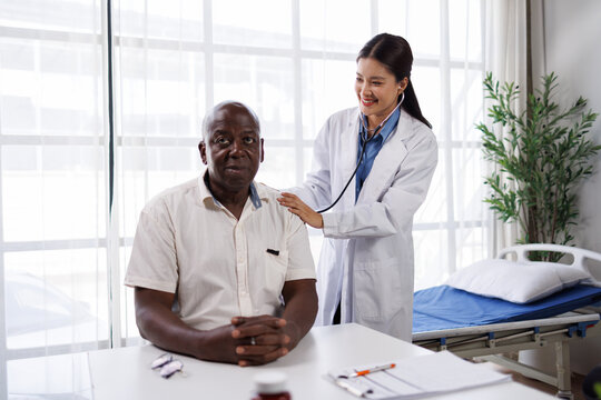 Female doctor in white coat performing a medical checkup on a smiling senior african american man using a stethoscope in a bright clinic room, focusing on his healthcare