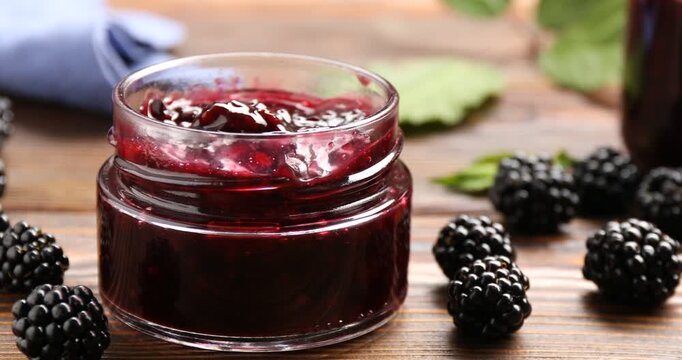 Delicious blackberry jam in glass jar, fresh berries and green leaves on wooden table, closeup. Camera moving right