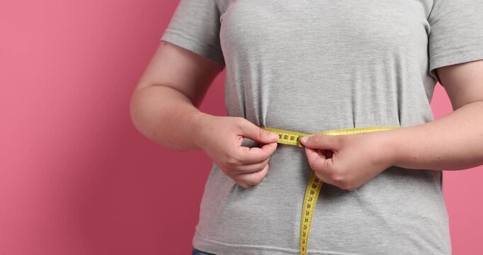 Weight loss. Overweight woman measuring her waist with tape on pink background, closeup