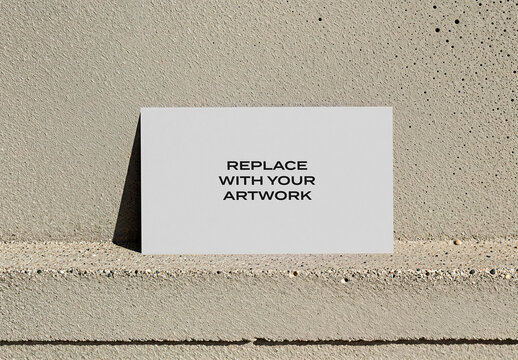Vertical business card mockup displayed on a concrete ledge against textured wall, realistic stationery presentation with natural sunlight and shadows in an outdoor urban environment