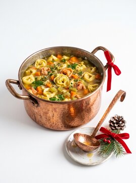 Traditional copper pot with tortellini in broth and red Christmas ribbon isolated on white