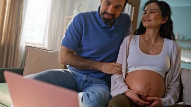 Expecting couple video call laptop home. Pregnant woman husband touch belly.