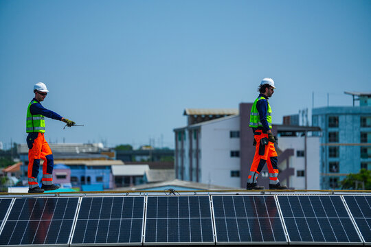 Two renewable energy specialists in hard hats and hi vis workwear deliberately walk the high ridge line of panels against the dense city skyline high on an industrial warehouse roof.