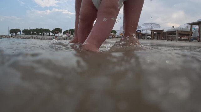 Parent standing in shallow ocean water, helping a baby taking first steps on a sandy beach during summer vacation