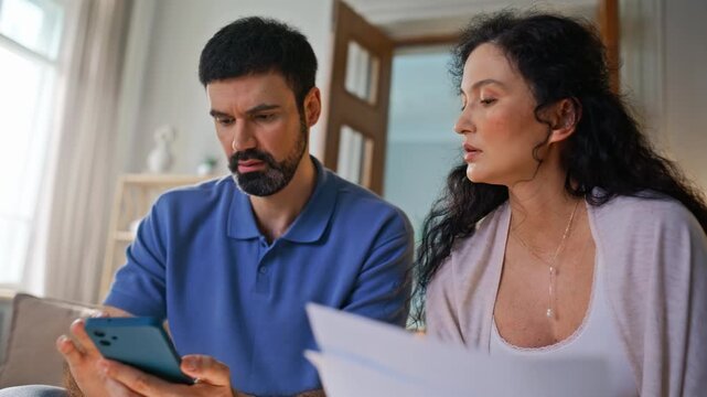Serious couple reviewing documents smartphone home. Stressed checking budget