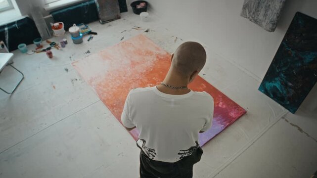 Caucasian young adult male artist walking around large abstract canvas on floor, evaluating fluid painting. Footage is perfect for creative industry marketing or art school promotions
