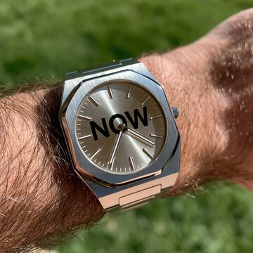 A watch marked NOW symbolizes the present moment, mindfulness, and the importance of using every breath and second of life meaningfully.