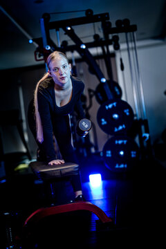 Women's strength and empowerment: a blonde Caucasian sportswoman training her back with a bent-over dumbbell row in a dimly lit gym, with determined concentration
