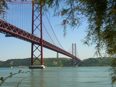 Red 25 de Abril Suspension Bridge Across Tagus River Framed by Tree Branches with Cristo Rei Sanctuary in Background, Landscape View in Lisbon, Infrastructure and Tourism Concept in Portugal