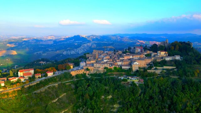 Mysterious aerial pedestal down shot from Santa Vittoria in Matenano with roads, buildings, trees, thickets, churches, belltower, clocktower, walls, surrounded by ravines and sky with various clouds