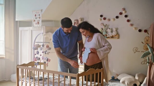 Couple preparing crib nursery. Pregnant wife showing baby bodysuit husband home.