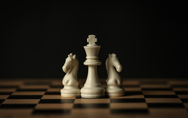 white chess pieces (king, knights) aligned on a wooden board, black background © geargodz