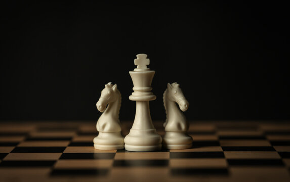 white chess pieces (king, knights) aligned on a wooden board, black background