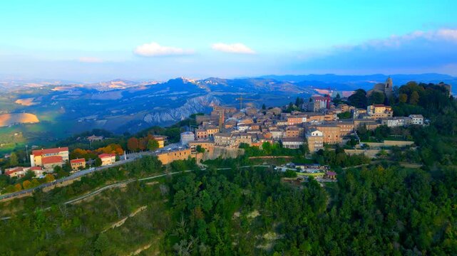 Verdant aerial push-in shot from Santa Vittoria in Matenano, the high-density town centre, a vintage human settlement surrounded by trees, thickets, ravines, typical Marche hillscapes, cloudy skies