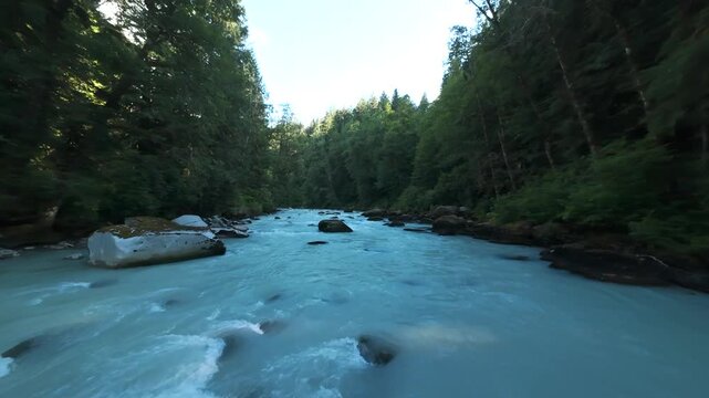 A fast-paced tracking aerial shot skims low against the current of a churning turquoise river, flying past mossy boulders as it follows a winding forest stream in the summer.