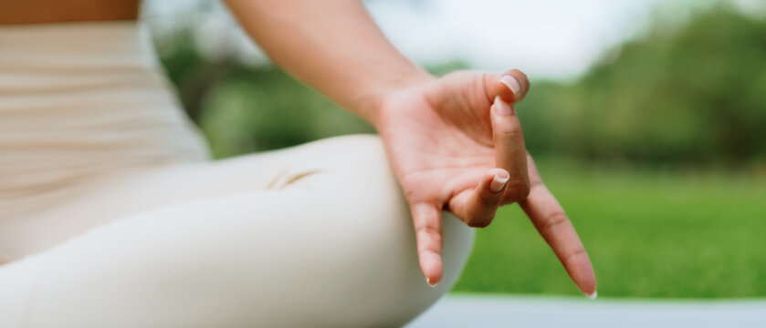 Asian woman meditating in a peaceful park setting, sitting cross-legged on green grass with trees in the background on a sunny day. Concept of mindfulness, self-care, and outdoor relaxation