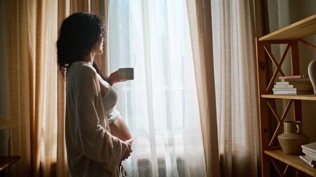 Pregnant woman drinking tea window. Expectant mother holding belly cozy home.
