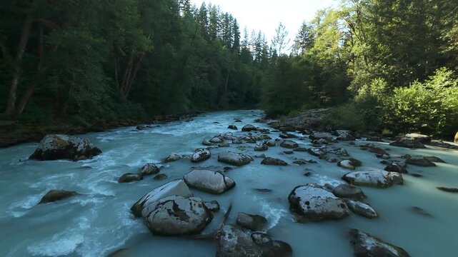 A descending aerial tracking shot low above a churning milky turquoise stream, gliding swiftly past rugged rocks and boulders scattered across a scenic forest riverbed.