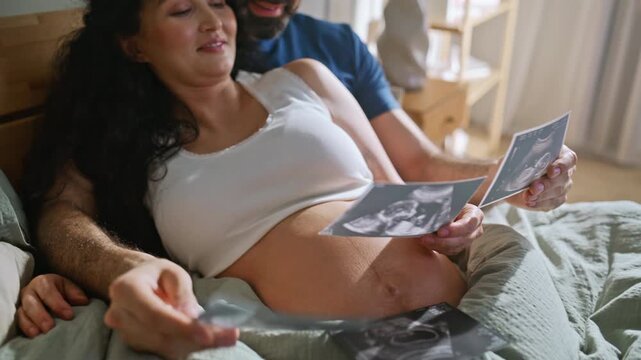 Pregnant woman husband looking sonogram photos bed. Expecting family bedroom.