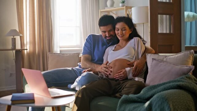 Pregnant wife husband touching belly watching laptop. Interracial couple home.