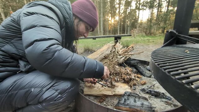 Woman making fire with a ferro rod at a campsite, igniting tinder with sparks to start a campfire for outdoor cooking and warmth during her adventure in the wilderness