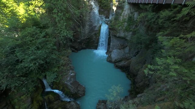 A high angle stationary aerial view looks down over thick green trees at a stunning mountain waterfall plunging into a wide, milky turquoise river canyon basin in the summer.