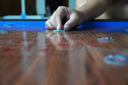 Close-up shot of a person's hand placing a green and white striker disc on a wooden carrom board surface. Focus on the finger and details of the plastic token. Traditional tabletop board game. karambo