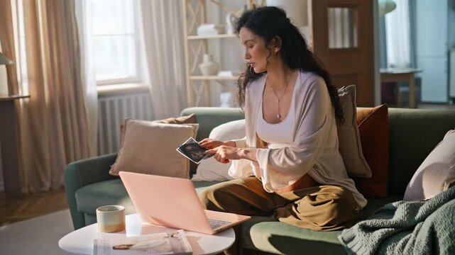 Mother-to-be showing ultrasound scan laptop video call. Pregnant woman sofa.