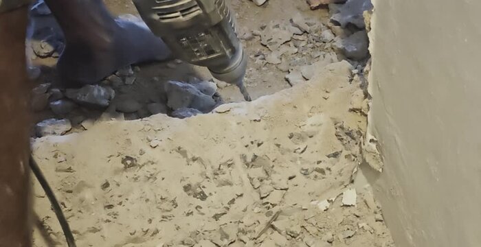 a worker using an electric rotary hammer drill to break down a concrete floor near an interior wall. Thick white dust clouds puff up as the drill bit chisels away at the hard concrete surface