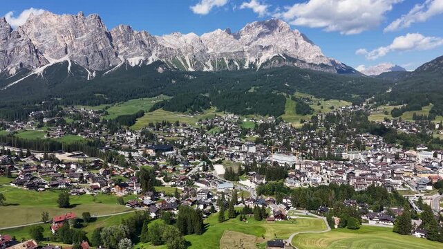 Aerial drone view over Cortina d'Ampezzo, showing the town surrounded by green alpine meadows and dense forests beneath the dramatic limestone peaks of the Tofane massif