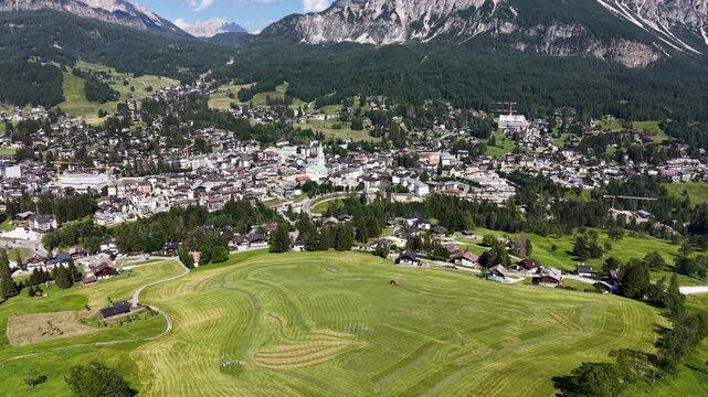 Aerial drone view over Cortina d'Ampezzo, revealing the town surrounded by mountain forests, alpine meadows, cultivated fields, and dramatic Dolomite peaks