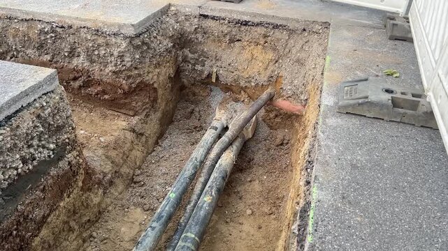 An open construction trench cuts along a residential street, exposing underground power cables and a sewer pipe in the layered soil. Groundwork and utility infrastructure in a German neighborhood.