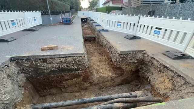 Slow forward camera move along a long open trench receding into the distance, exposing the same underground power cables and sewer pipe in the soil. Groundwork and utility infrastructure, German neigh