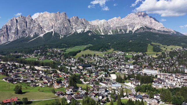 Aerial drone view across Cortina d'Ampezzo, with the town's rooftops and alpine meadows set against the imposing limestone cliffs of the Tofane massif beneath a clear summer sky