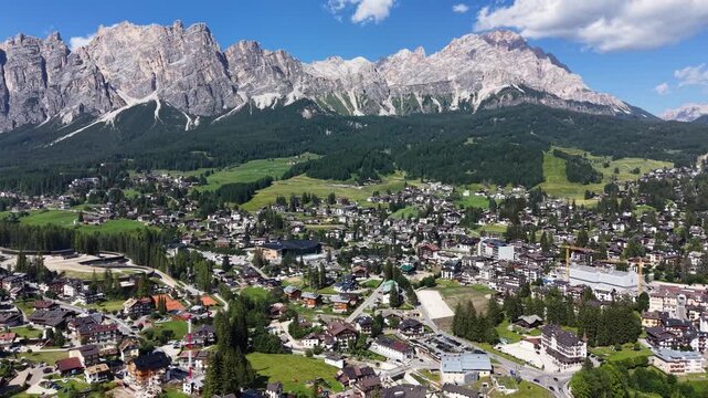Aerial drone view over Cortina d'Ampezzo, with the extensive townscape and green alpine meadows set beneath the dramatic limestone peaks of the Tofane massif