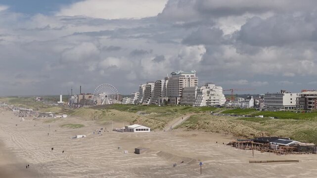 Noordwijk beach stretches below dunes and a Ferris wheel, with hotels and apartment towers lining the coastal resort near Palaceplein, South Holland, Netherlands, orbiting establishing shot.