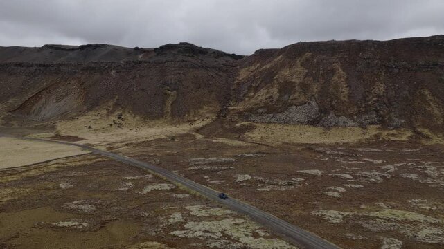 Car travels along Road 42 through barren volcanic terrain near Vatnsskar&eth; on the Reykjanes Peninsula, Southern Peninsula, Iceland, with dark ridges surrounding the roadway, drone tracking shot.