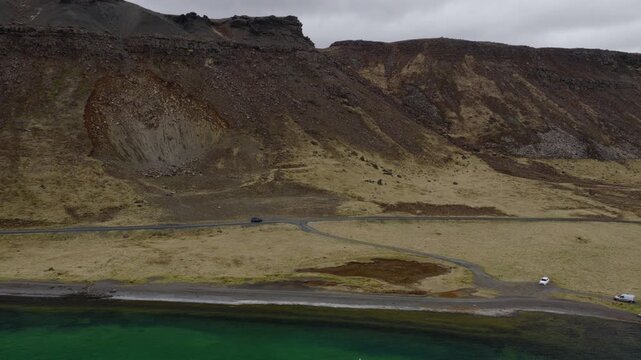 Car travels along Road 42 beside Kleifarvatn on the Reykjanes Peninsula, with rugged volcanic slopes rising above the lake in Grindavikurbaer, Iceland, drone tracking shot.