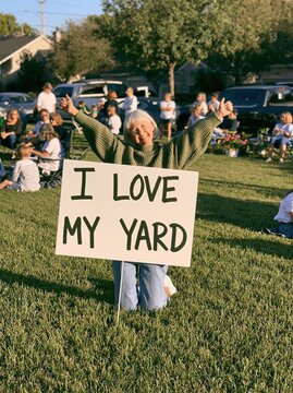 Joyful Senior Woman Holding "I Love My Yard" Sign at Outdoor Gathering on Sunny Day