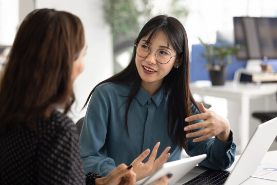 Two women working together at desk with laptop, using tablet, talking, collaborating on project, brainstorming, engaged in focused teamwork in office. Female manager consulting client during meeting