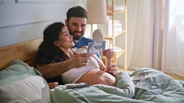 Couple viewing ultrasound scan bed. Pregnant woman husband hold sonogram home.