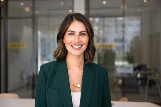 Head shot of young Mexican businesswoman dressed in elegant jacket, smiling, looking at camera standing alone in contemporary meeting room, posing for photo. Office portrait of female CEO or executive