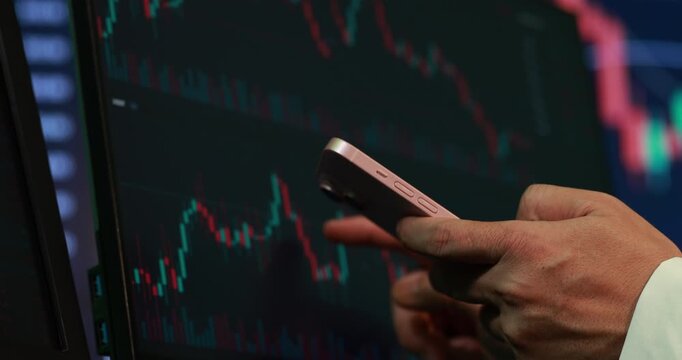 Hand holding a smartphone in front of a stock market chart screen with falling and rising prices